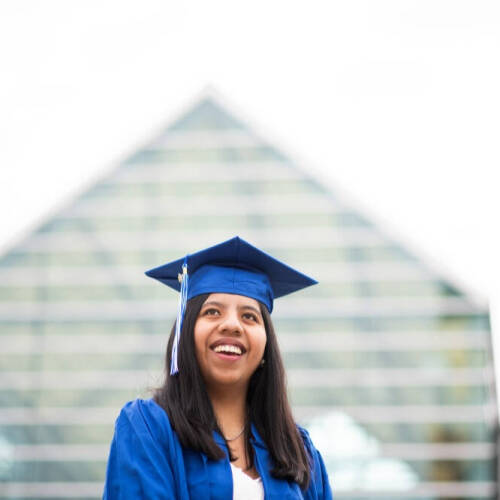 A young woman wearing a blue graduation cap and gown smiles as she stands in front of a modern glass‑and‑steel building that appears campus‑like.